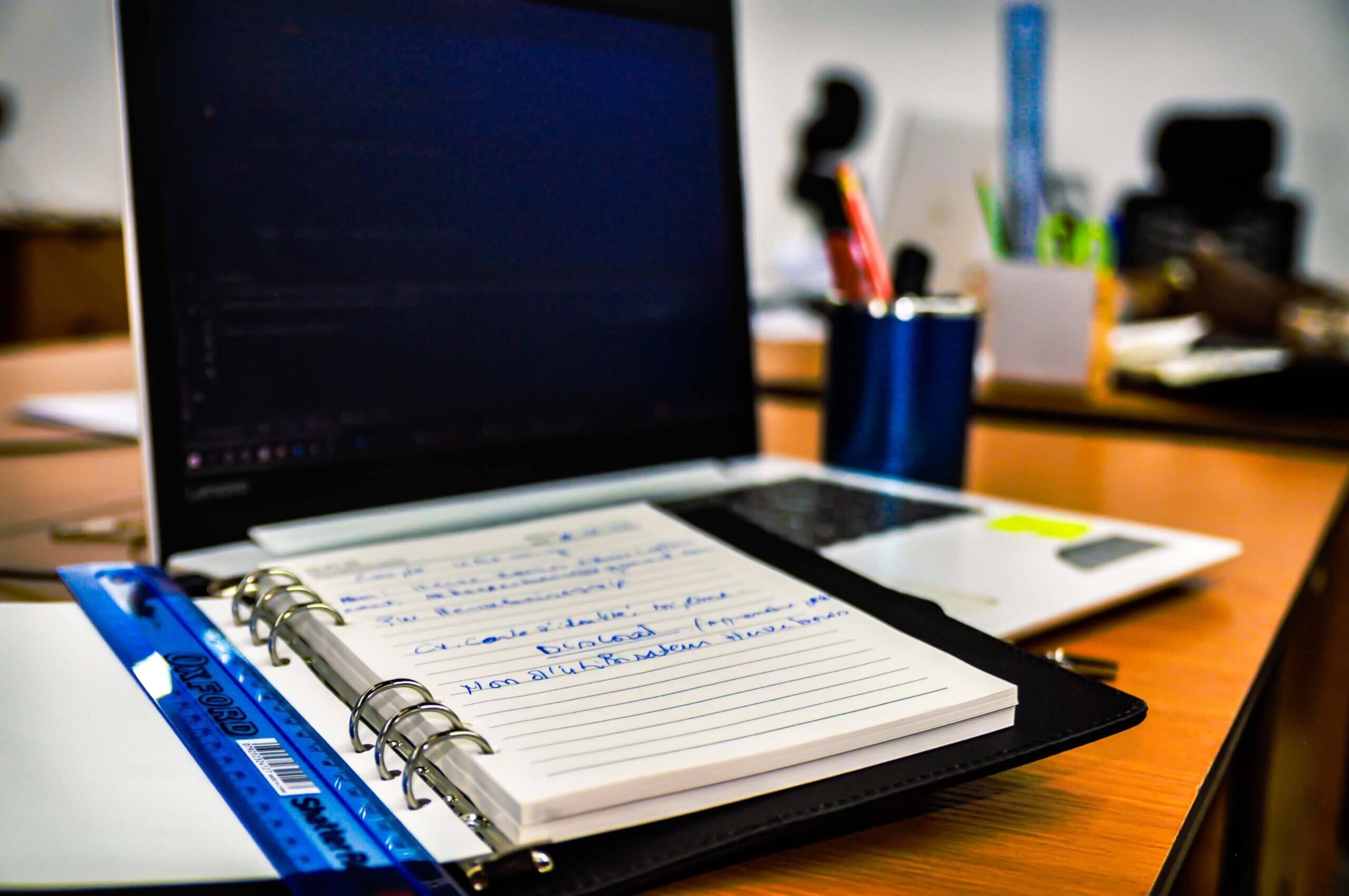 Open spiral notebook with blue-ink notes on a wooden desk in front of a laptop.