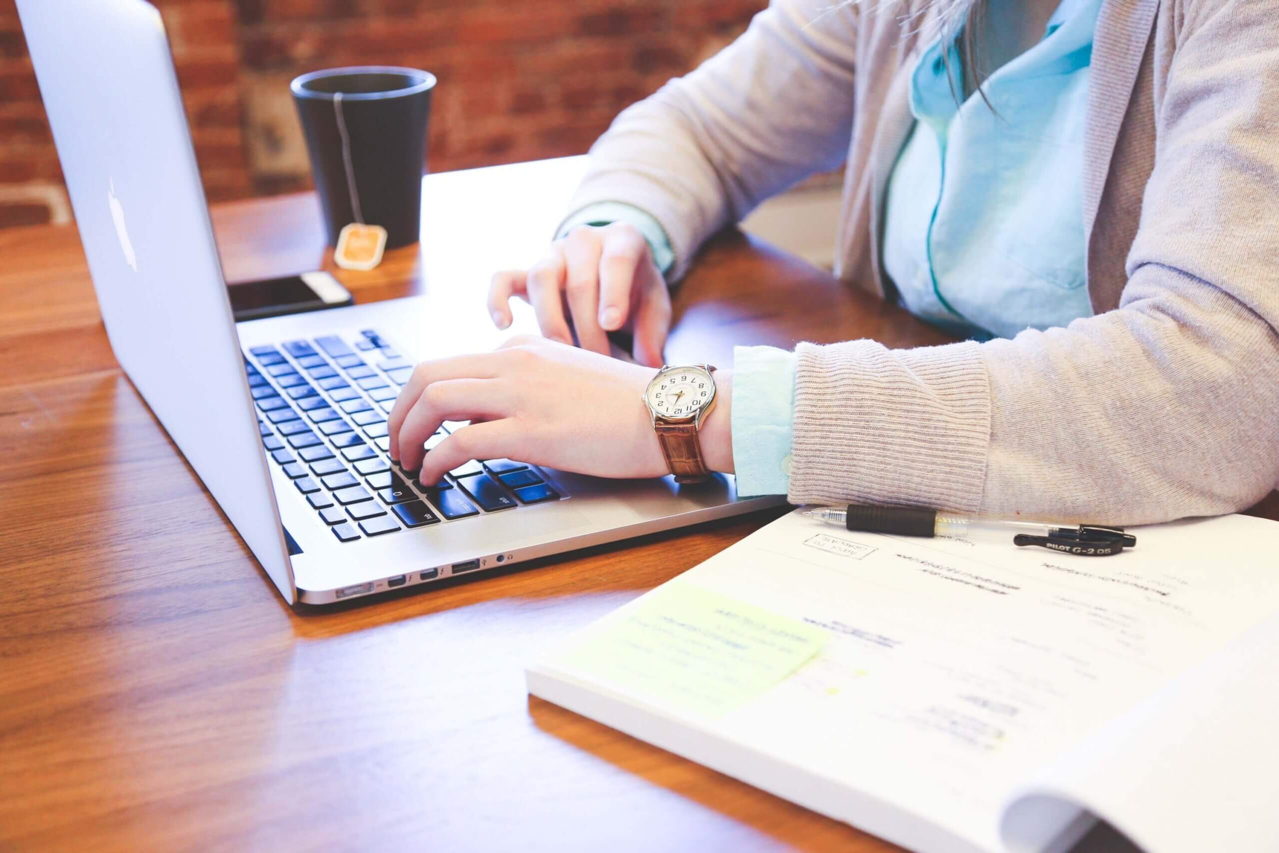 Person typing on a silver laptop at a wooden table, with a coffee cup and a notebook and markers nearby.